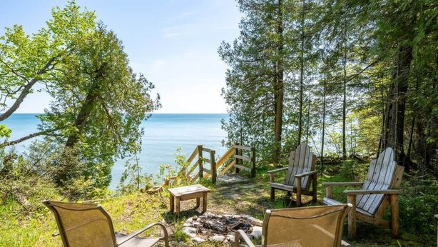 A serene outdoor seating area with Adirondack chairs and a fire pit, overlooking a calm lake and dense forest. - Lake Michigan Cabin · Private Shoreline (Sturgeon Bay)
