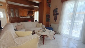 Un salon avec un grand canapé beige, une table basse et un lustre. On y trouve également un coin cuisine avec des meubles en bois et une fenêtre agrémentée de rideaux.