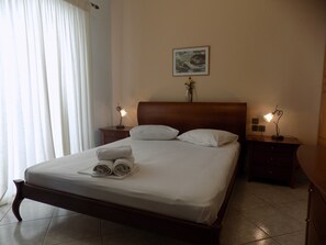 A neatly made bed with white linens and rolled towels, two bedside tables with lamps, a framed picture on the wall, and a window with white curtains. - Beautiful Modern apt in Skiathos (Skiathos)