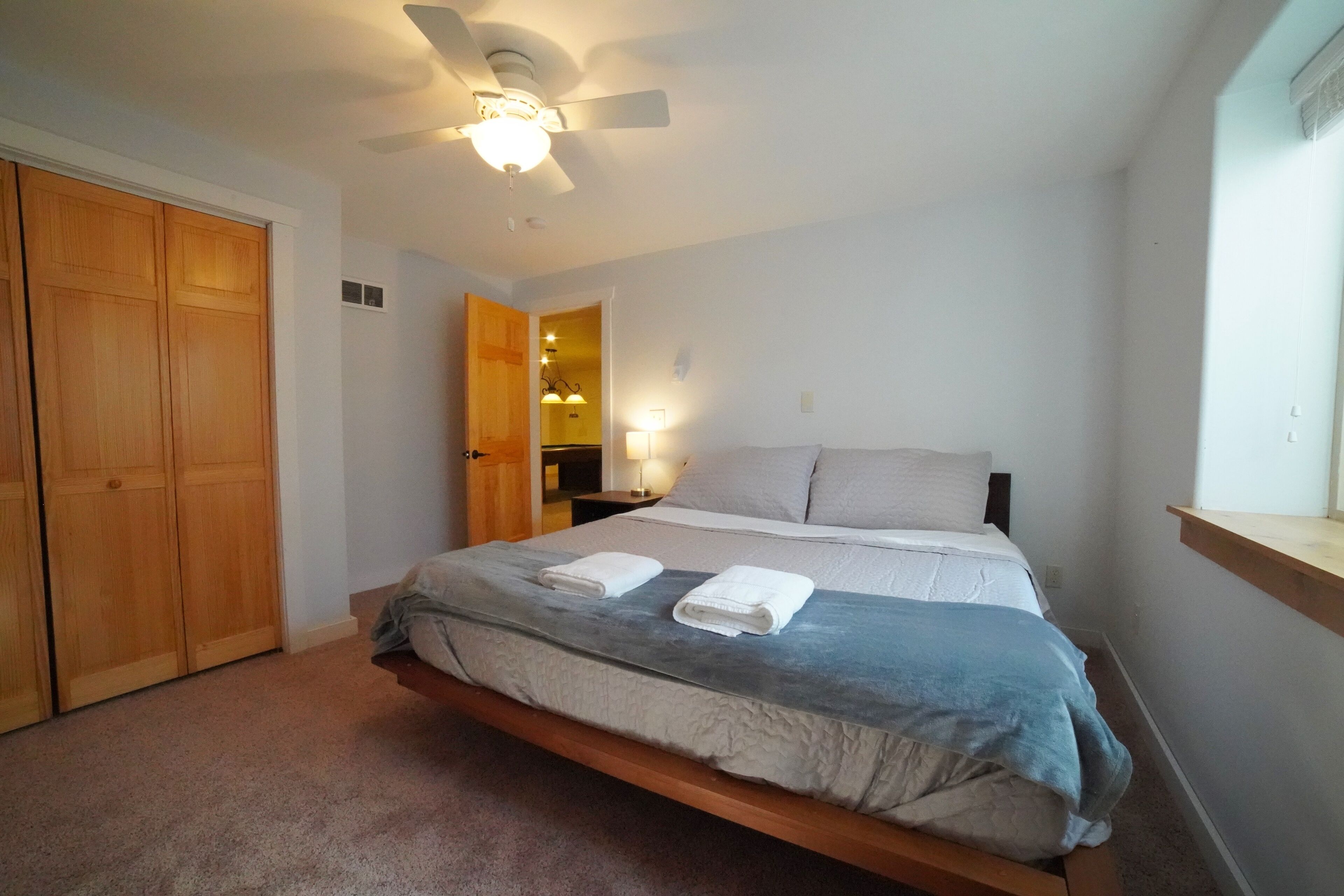 Quarto com cama, ventoinha no teto, guarda-roupa de madeira e mesa de cabeceira com abajur.