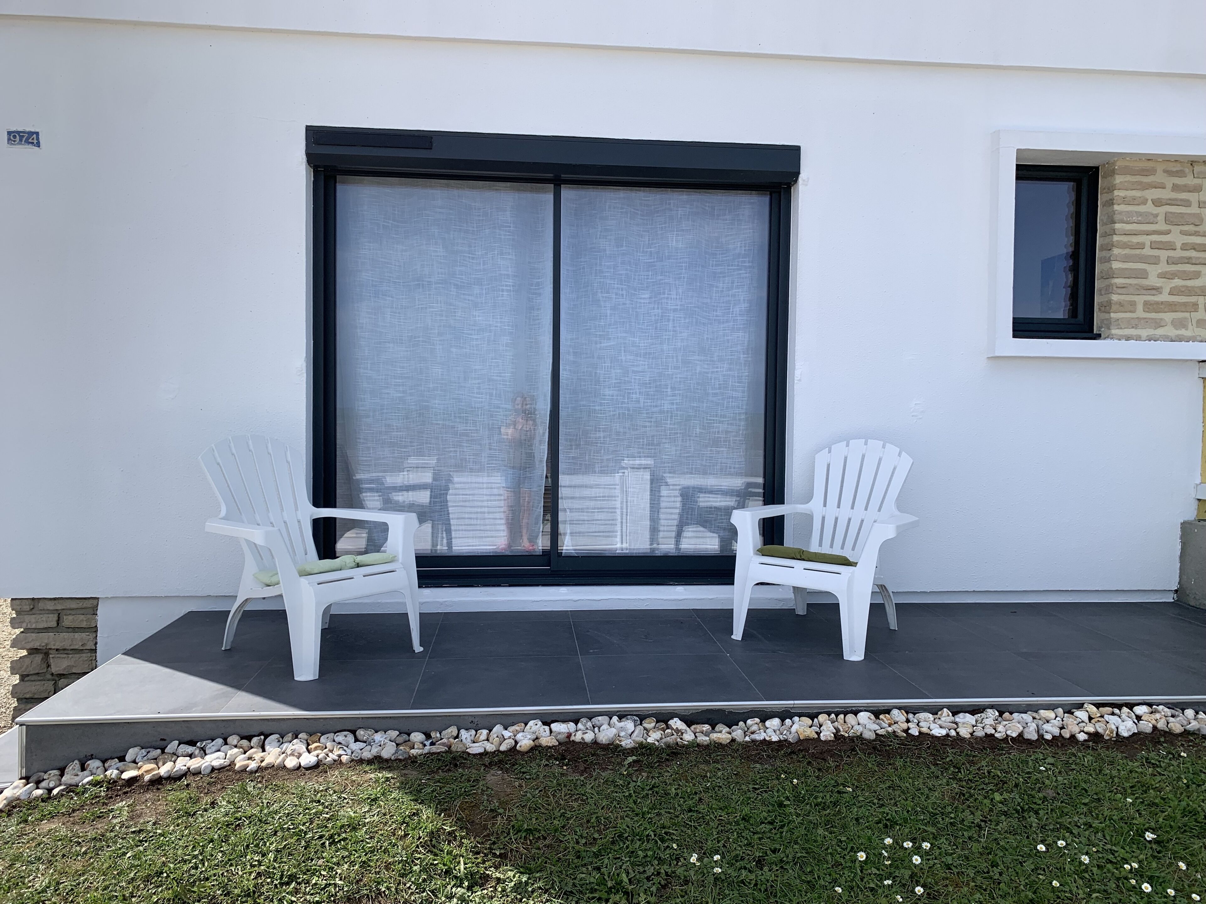A patio with two white chairs, a sliding glass door, and a small window with a brick wall.