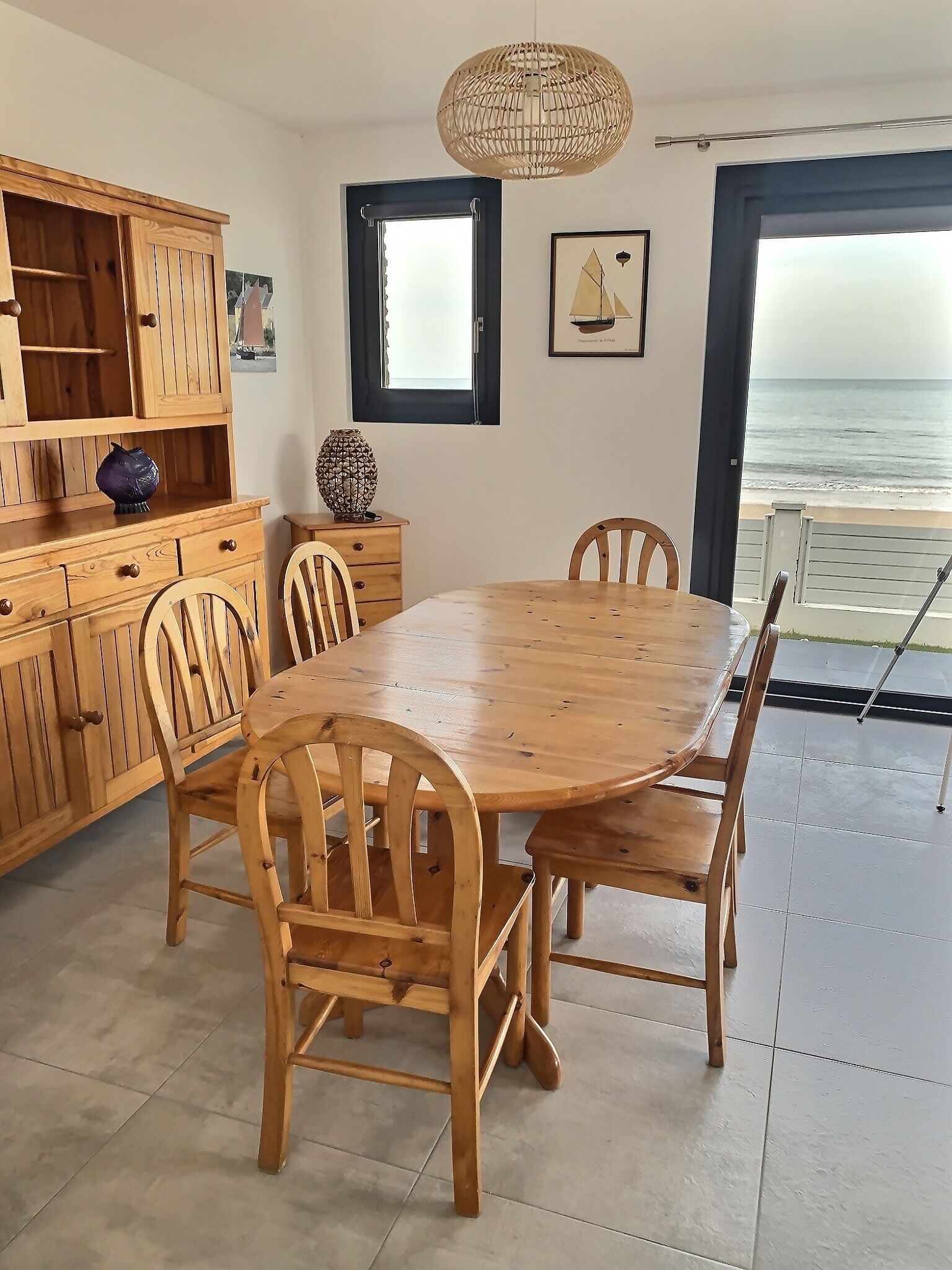 Uma mesa de jantar de madeira com cadeiras, um armário de madeira com gavetas, um lustre pendente, uma janela com vista para o oceano e uma moldura com um quadro na parede.