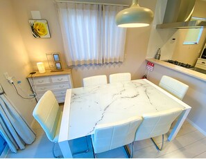 A dining area with a marble table and white chairs, a chandelier, a stove, and a window with curtains.