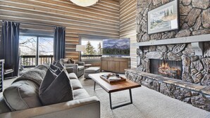 A cozy living room with a stone fireplace, a large TV, and a view of mountains through the windows.