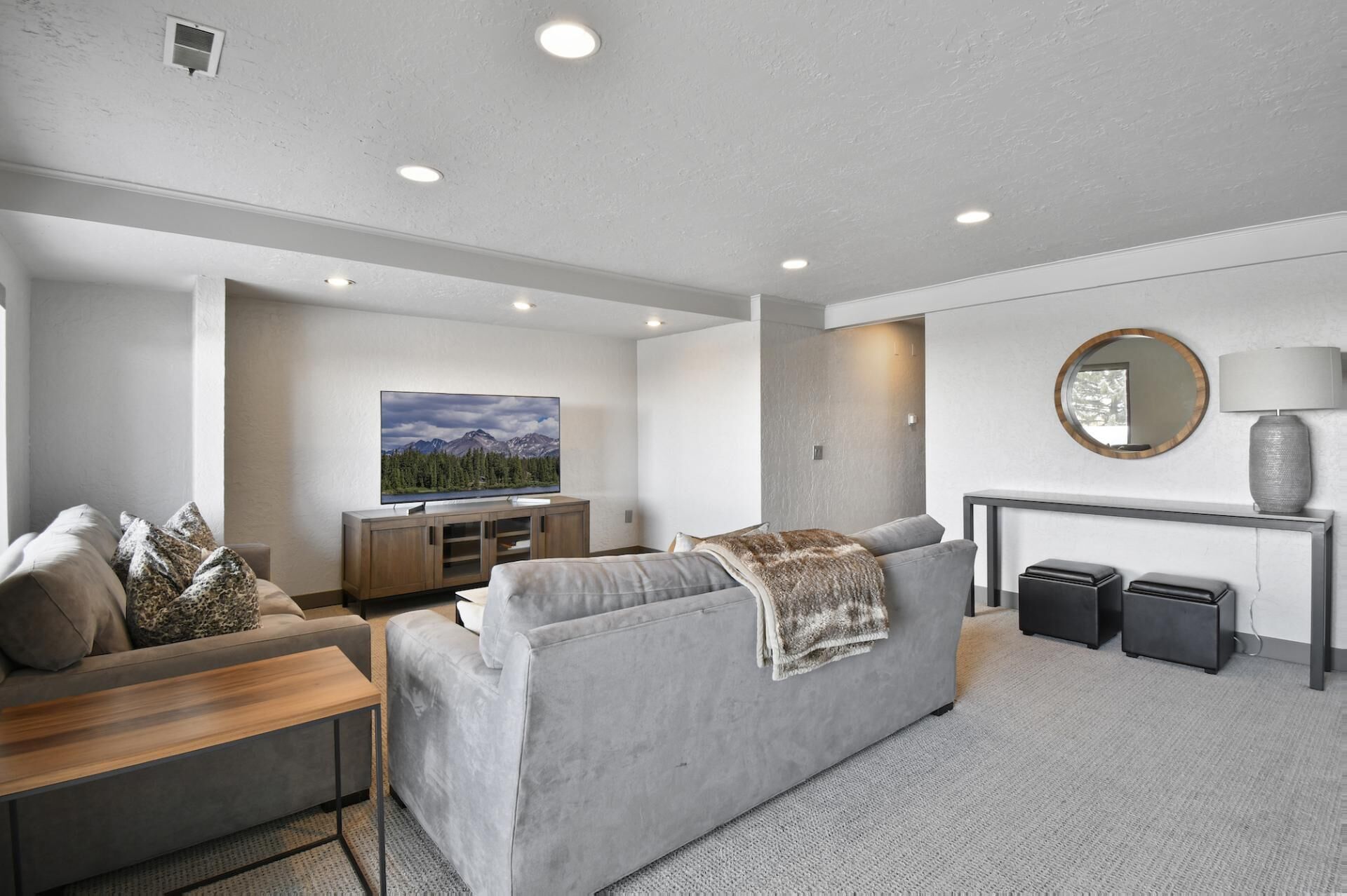 A modern living room with a grey sofa, a wooden coffee table, and a flat-screen TV on a wooden stand. There is a round mirror on a console table and a lamp on the console table.