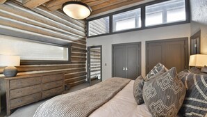 A bedroom with a log wall, a large bed with patterned pillows, a dresser, and a window above the bed.