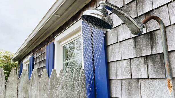 Ein Metall-Duschkopf an der Außenwand eines Hauses, mit Wasser, das in einen blauen Regenfall fließt.