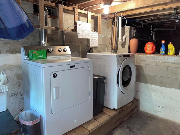 A laundry area with a washing machine and dryer, a trash bin, and cleaning supplies on a shelf.