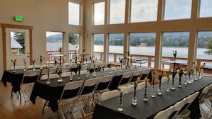 A conference room with tables set for a meeting, large windows with a lake view, and an exit sign on the wall. - Grewingk Suite - Homer Seaplane Base (Homer)