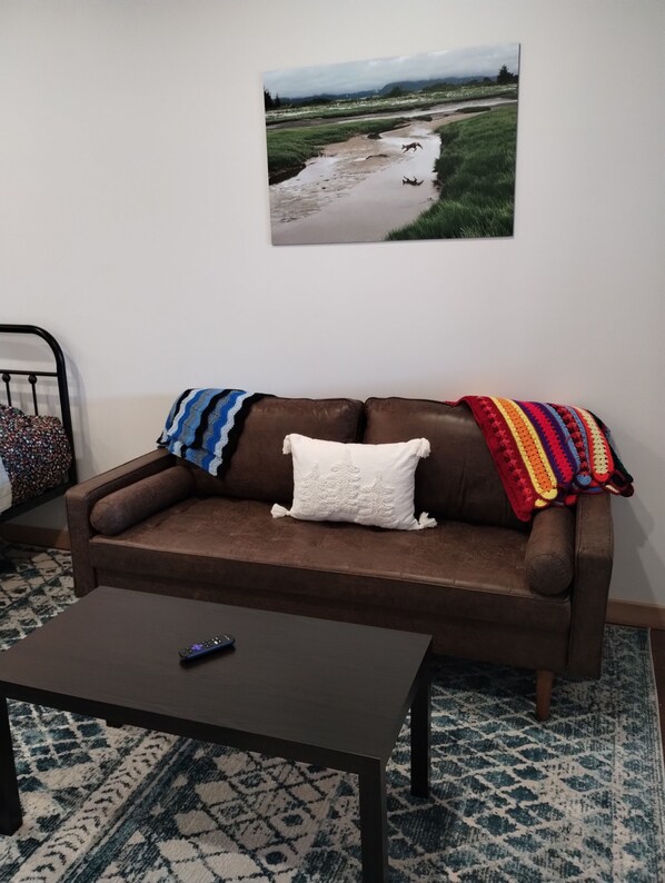 A brown sofa with a white pillow and a colorful blanket, a remote control on a dark coffee table, and a framed landscape painting on the wall. - Leisure Suite - Homer Seaplane Base (Homer)