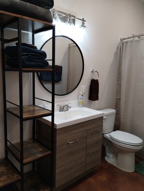 A bathroom with a wooden vanity, a round mirror, a toilet, and a shower curtain. - Leisure Suite - Homer Seaplane Base (Homer)
