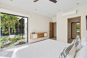 A bedroom with a large bed, a wooden bedside table, a built-in bench, and a view of a garden through a sliding door.