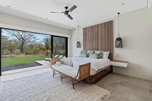 A bedroom with a large bed, a ceiling fan, a view of the outdoors, and a hanging light fixture.