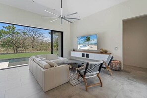 A modern living room with a large window, a white sofa, a dining table with chairs, and a flat-screen TV mounted on the wall.