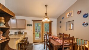 A kitchen with wooden cabinets, a dining table with chairs, a hanging light fixture, and a view of the outdoors through a glass door. - Pet-friendly cabin in the pines, 2 decks w/grill, <10 to skiing (Angel Fire)