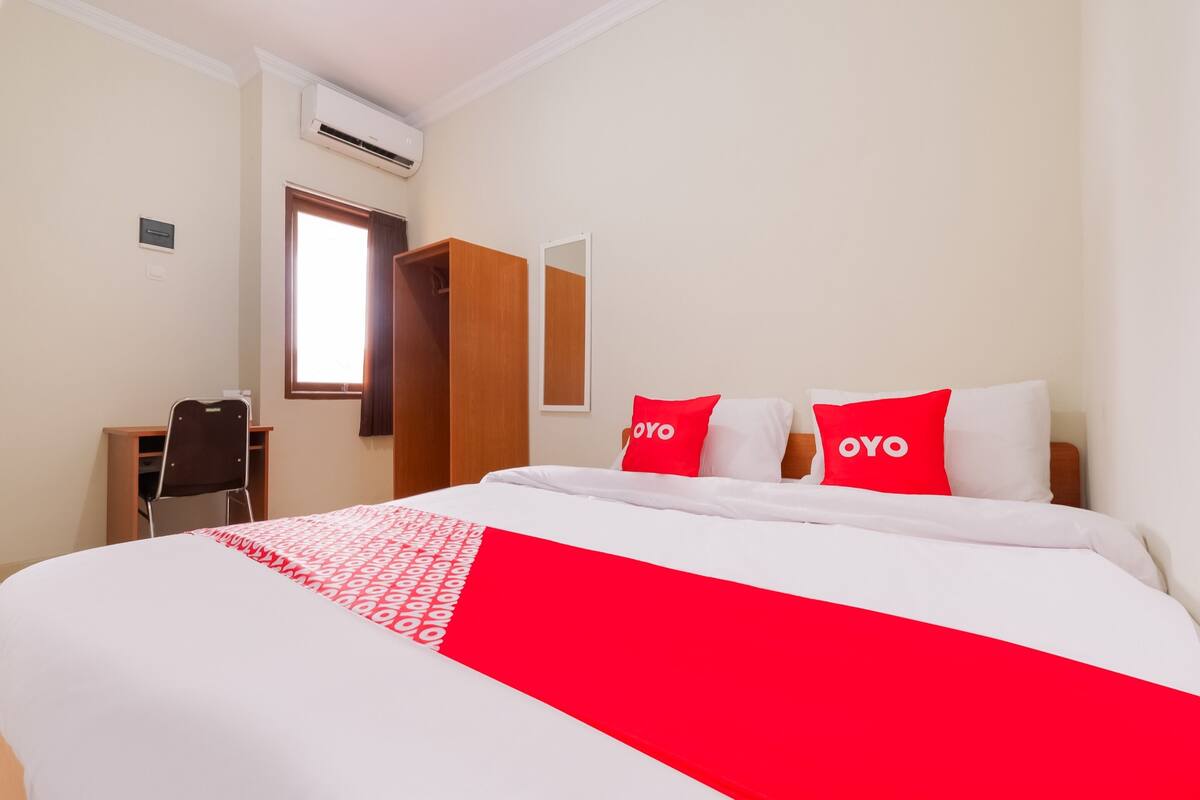 A hotel room with a bed, two red pillows with the word "OYO" on them, a wooden wardrobe, a desk with a chair, and a window with a view.