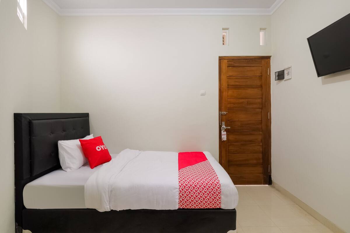 A single bed with a black headboard, white bedding, and a red pillow. A wooden door is visible in the background.