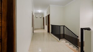 Un couloir propre et bien éclairé, avec une porte en bois sur la gauche, un escalier avec des rampes noires et un mur blanc avec une porte au fond.