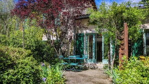 Een tuinpad dat naar een gebouw leidt, met een groene deur, omgeven door weelderige begroeiing en een houten constructie.