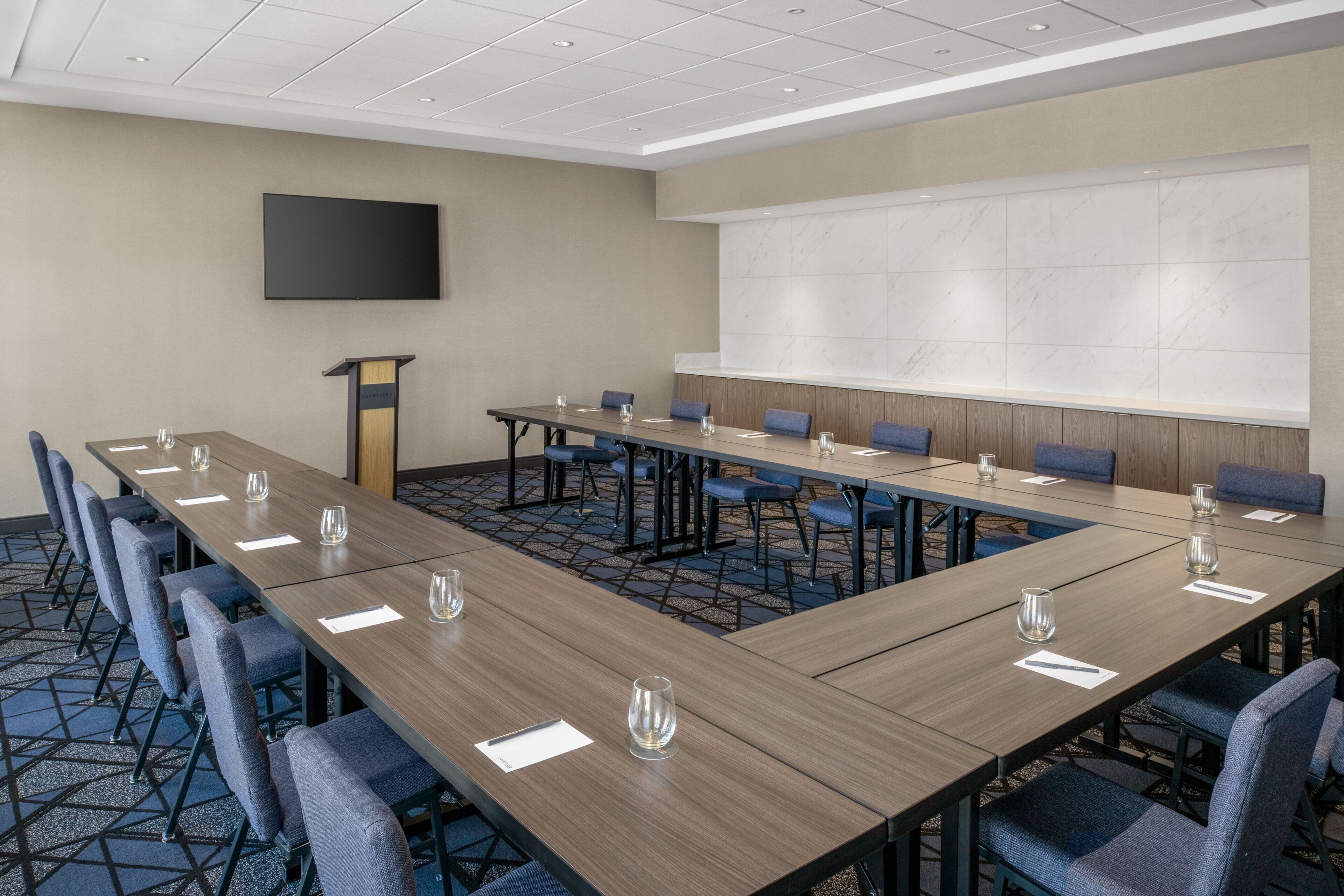 A conference room with a long table, blue chairs, a podium, and a large screen.