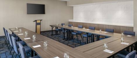 A conference room with a long table, blue chairs, a podium, and a large screen.