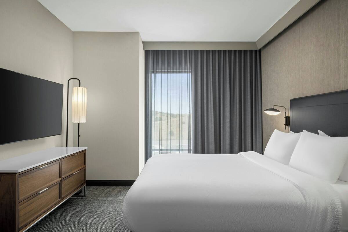 A hotel room with a large bed, a flat-screen TV, a dresser, and a window with curtains.