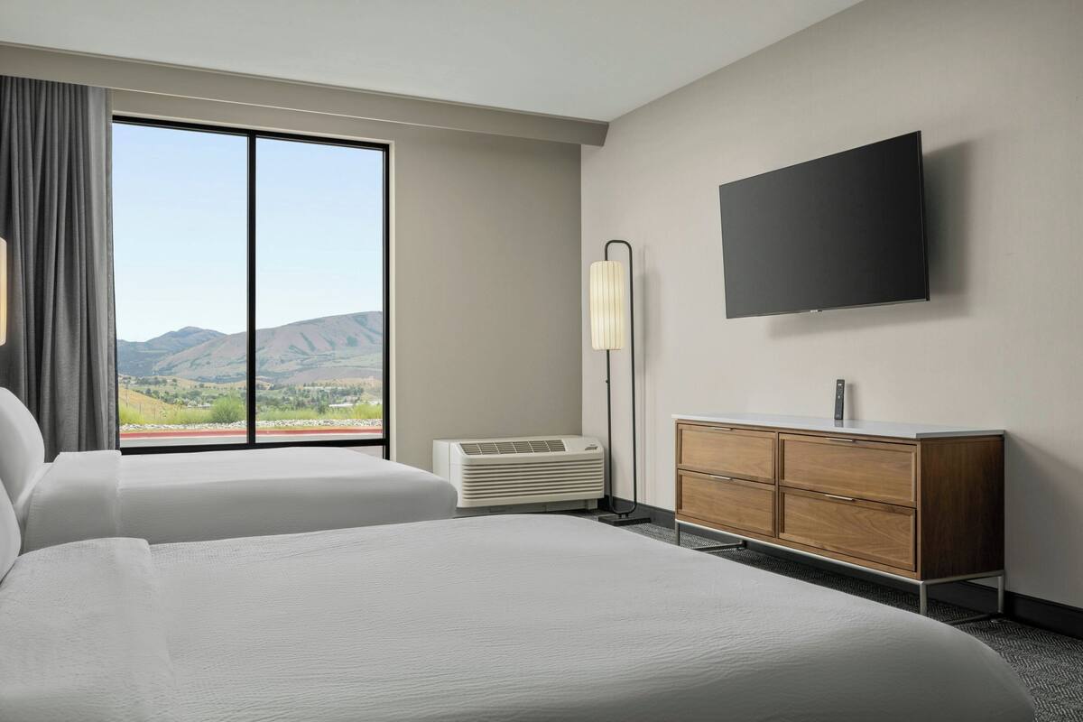 A hotel room with two beds, a flat-screen TV, a bedside table, a lamp, and a view of mountains.