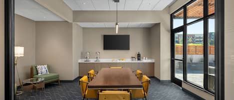 A modern conference room with a long table, yellow chairs, a sofa, a TV, and a kitchen area with a large window.