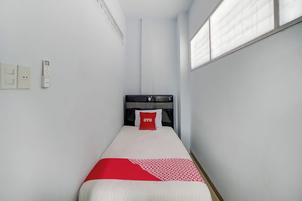 Ein schmales Hotelzimmer mit einem Einzelbett, einem Nachttisch, einer Steckdose und einem Fenster mit Jalousien.