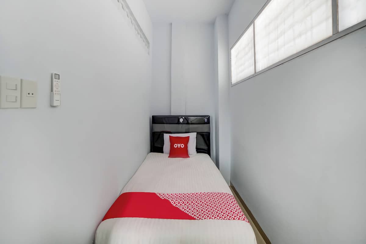 A narrow hotel room with a single bed, a bedside table, a power outlet, and a window with blinds.