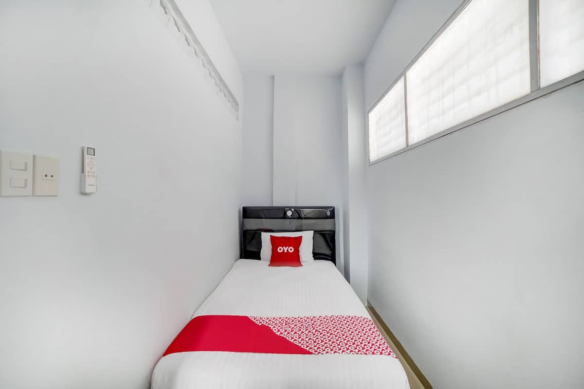 A narrow hotel room with a single bed, a red pillow, and a white bedspread.