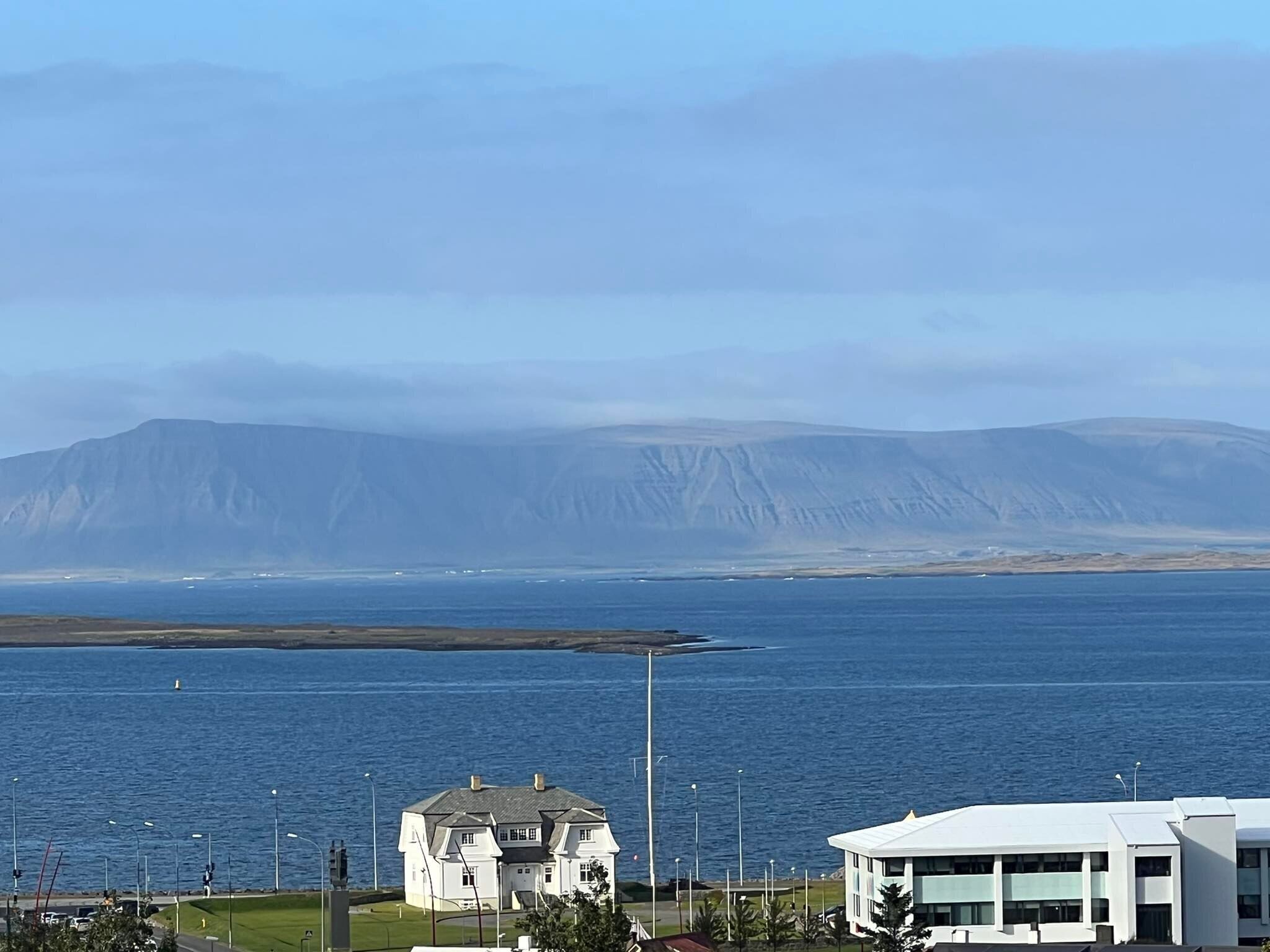 A coastal view with a large mountain in the background, a white building with a green lawn in the foreground, and a modern white building with a flat roof.