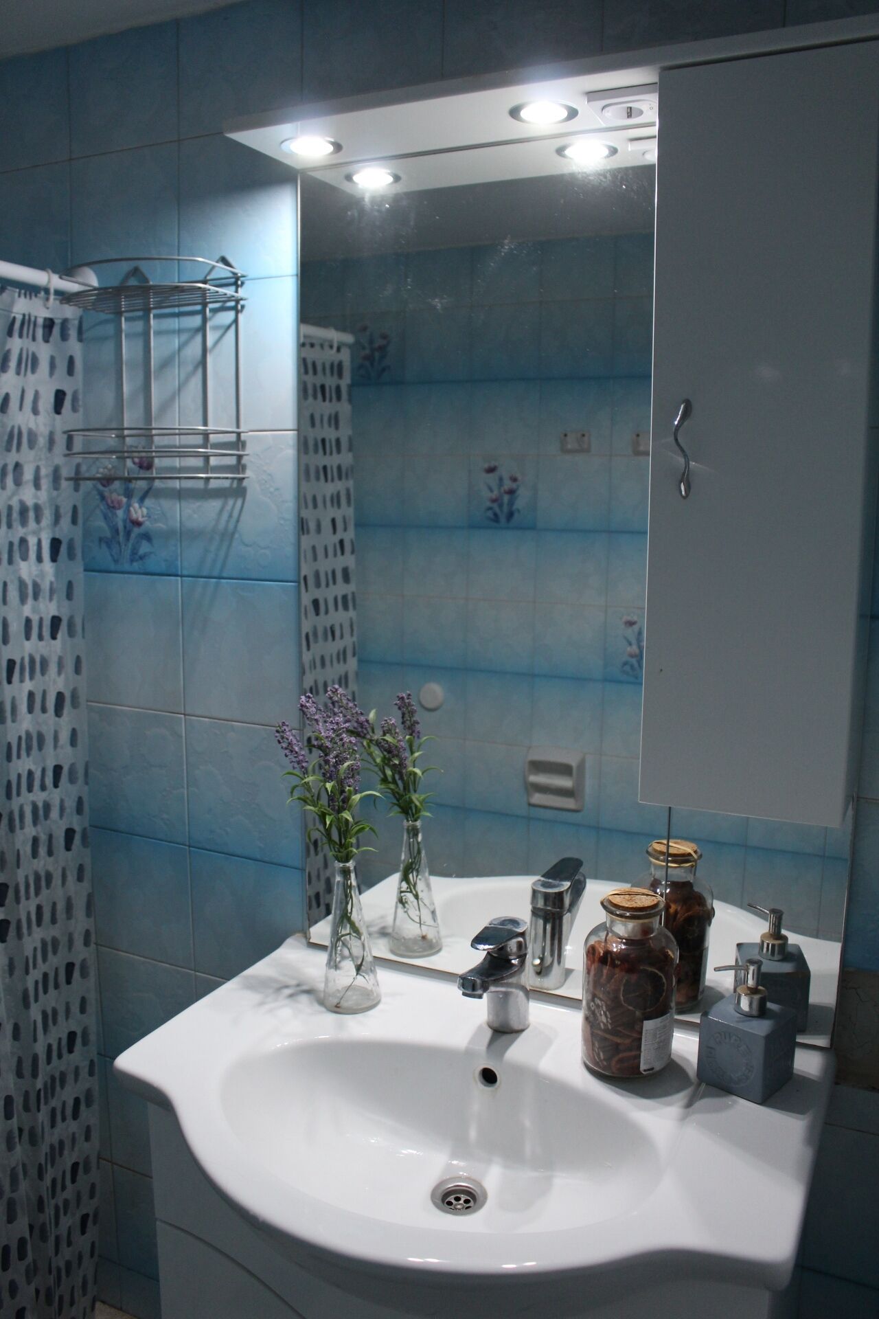A bathroom with a white sink, a mirror, and a blue tiled wall. There are two glass vases with flowers on the sink counter.