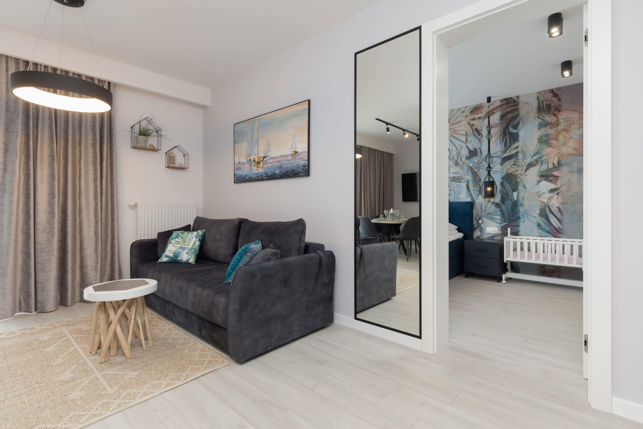 Ein modernes Wohnzimmer mit einem grauen Sofa, einem kleinen Holztischchen, einem großen Spiegel und einem Gemälde mit Segelbooten.
