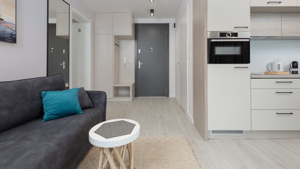 A modern apartment with a grey sofa, a small round table, a kitchen with white cabinets, and a grey door. - Cozy Apartment | Close to Beach and Lake, Parking. (Mielno)