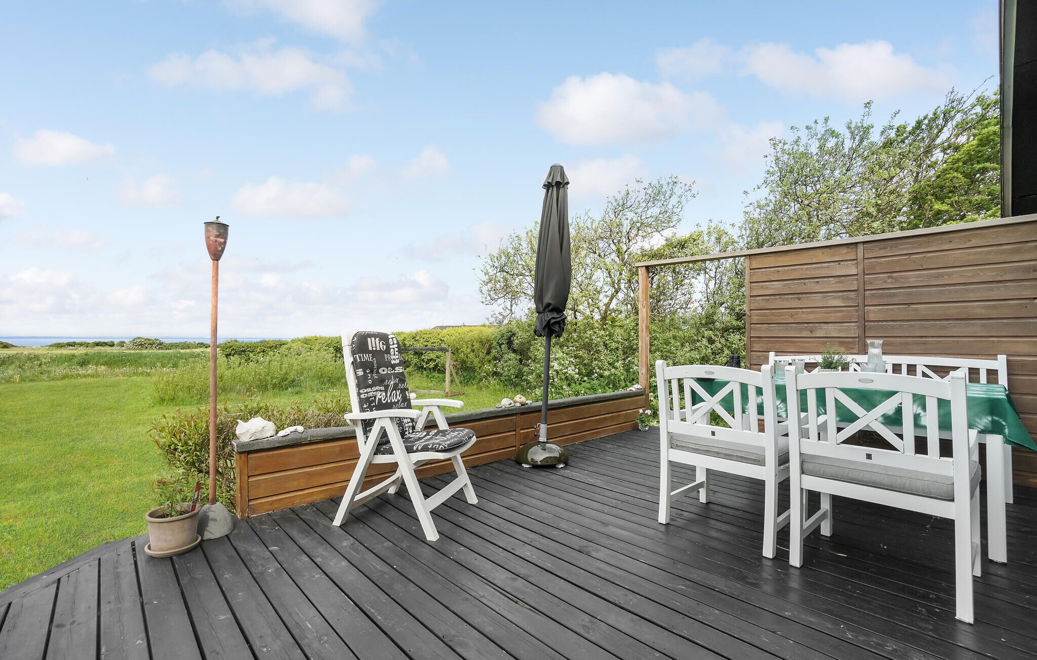 A wooden deck with outdoor furniture, including a white chair and a table with a green cushion, overlooking a grassy area and a body of water.