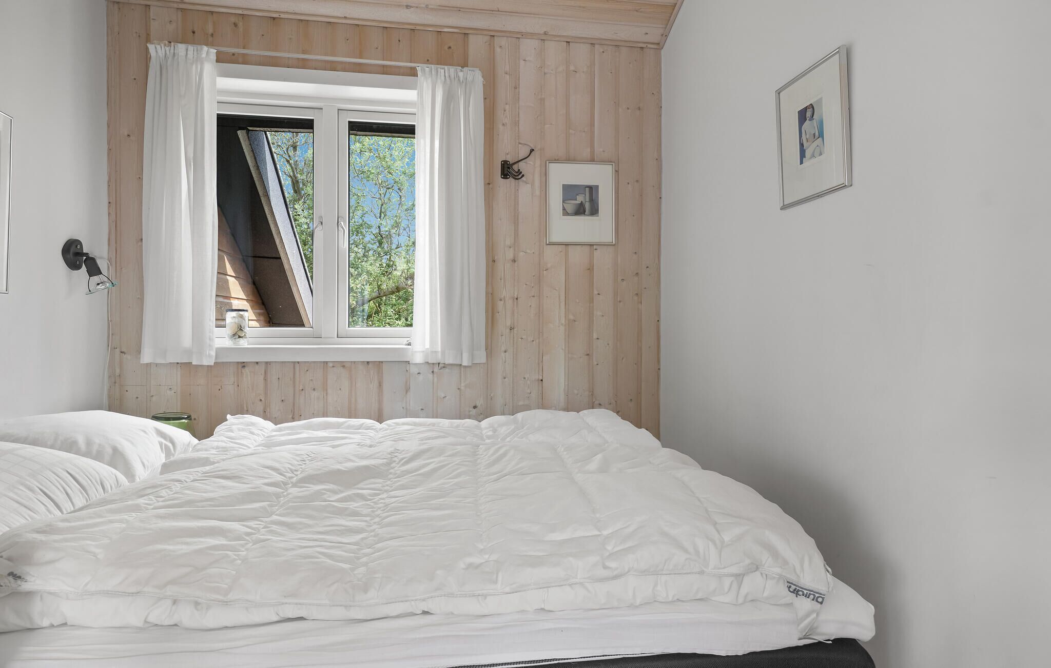Et soveværelse med en seng, et vindue med gardiner, en billedramme og en lampe på væggen.