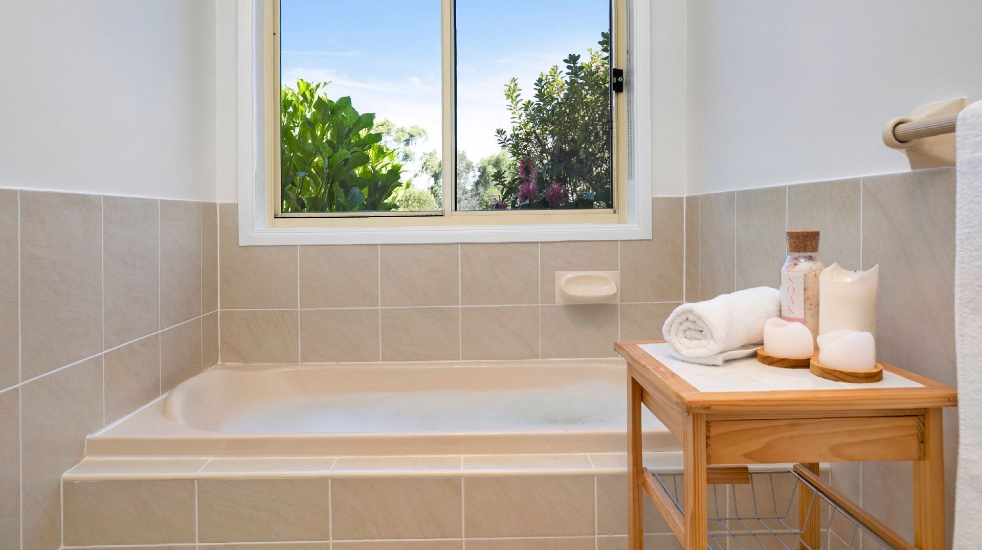 Ein Badezimmer mit einer Badewanne, einem Fenster mit Blick auf Grünflächen und einem kleinen Holztisch mit Toilettenartikeln.