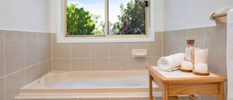 Ein Badezimmer mit einer Badewanne, einem Fenster mit Blick auf Grünflächen und einem kleinen Holztisch mit Toilettenartikeln.