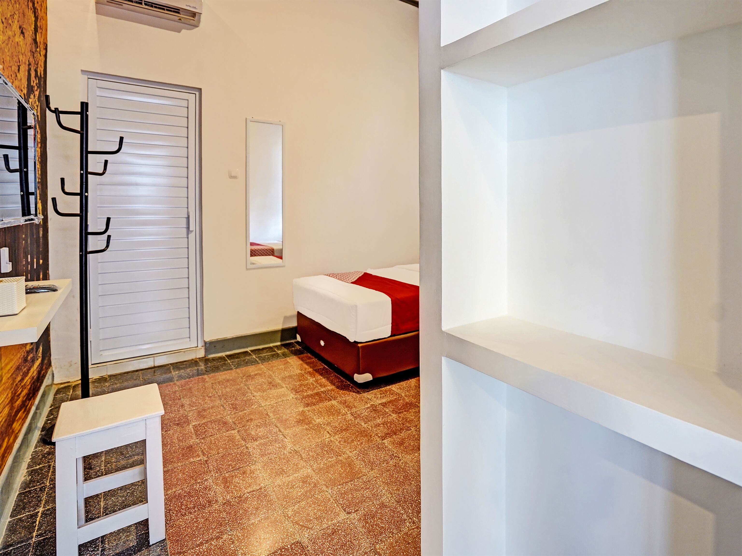 Una habitación de hotel con una cama, un taburete blanco pequeño, un espejo y una puerta blanca con picaporte.