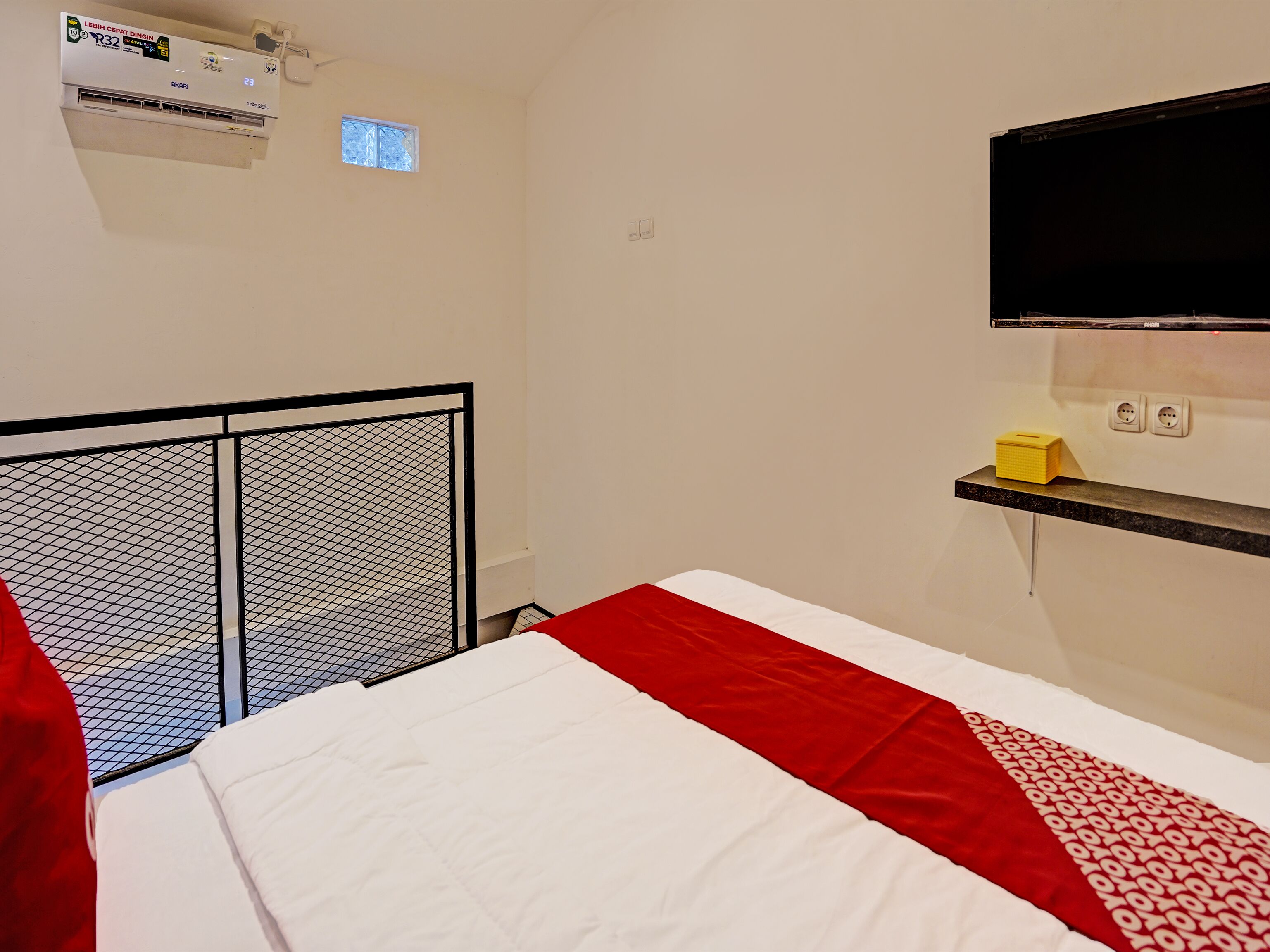 Ein Hotelzimmer mit Bett, Wandfernseher, kleiner Ablagefläche mit einem gelben Gegenstand und Klimaanlage.