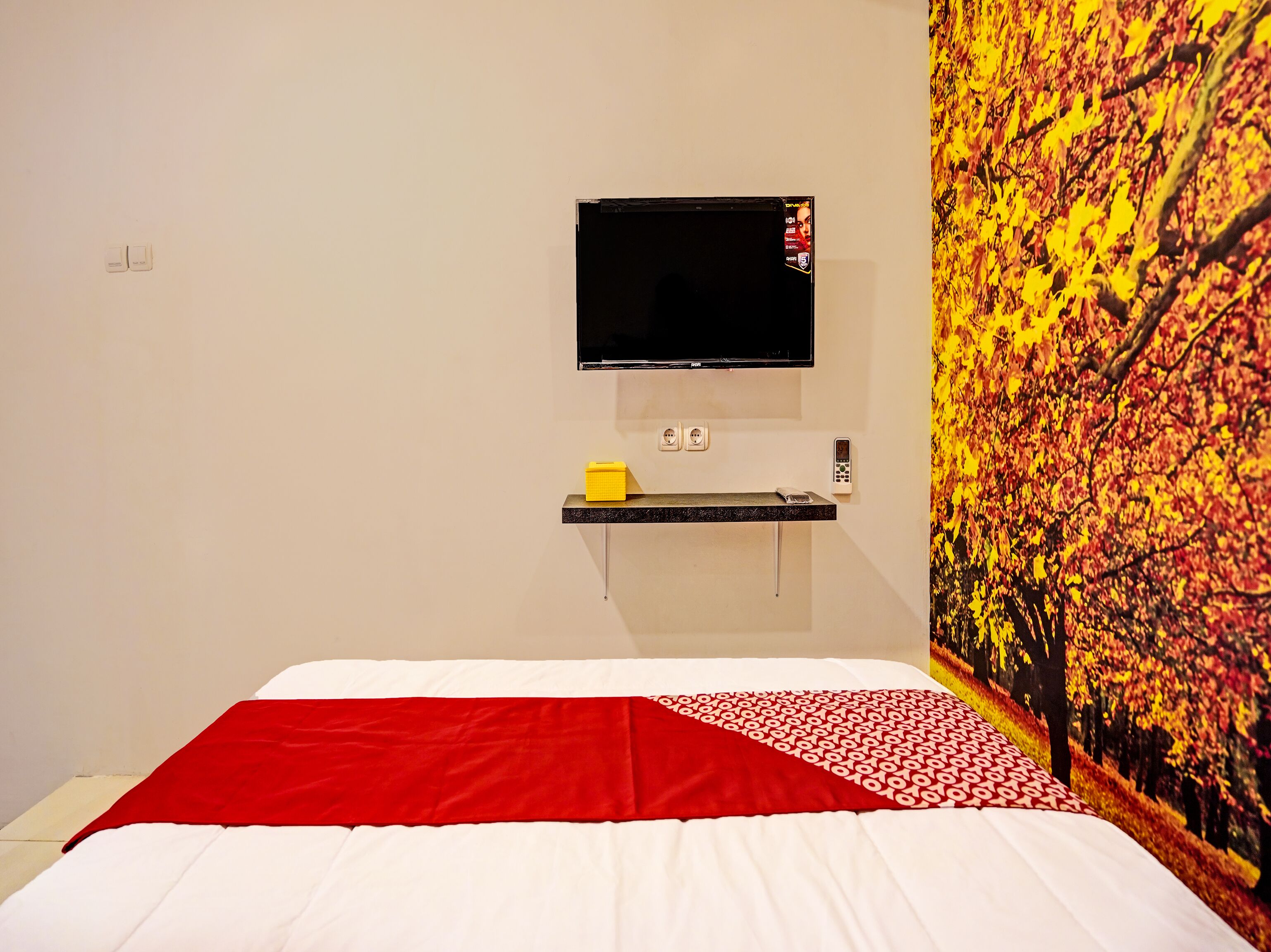 A hotel room with a bed, a mounted TV, a small shelf with a phone and a yellow box, and a wall with a tree mural.