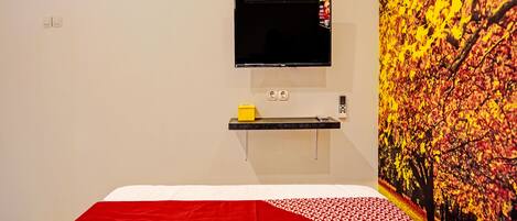 A hotel room with a bed, a mounted TV, a small shelf with a phone and a yellow box, and a wall with a tree mural.