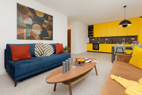 Una sala de estar moderna con un sofá azul, una mesa de centro de madera y una cocina amarilla al fondo.