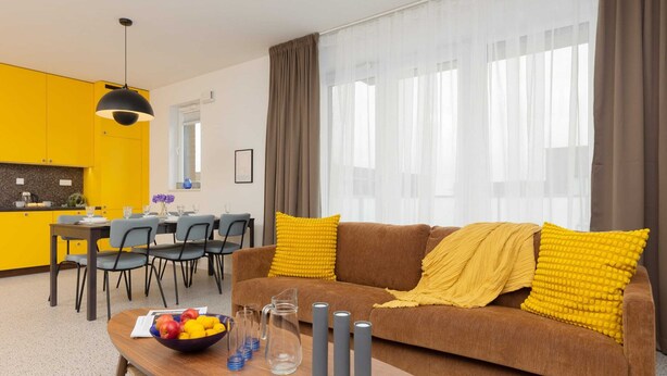A modern living room with a brown sofa, yellow cushions, and a wooden coffee table. There is a dining area with a table and chairs in the background.