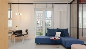 Un salon moderne avec un canapé bleu, une petite table sur laquelle repose un magazine, un fauteuil et une fenêtre donnant sur des immeubles.
