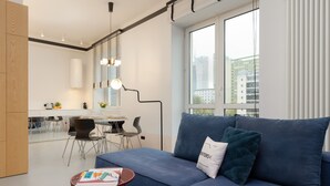 Un salon moderne avec un canapé bleu, une petite table ronde sur laquelle repose un magazine, et une vue sur la ville à travers de grandes fenêtres.