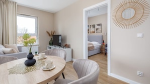 A modern living room with a round wooden table, a sofa, and a TV. There is a bed in the background with a view of the outdoors. - Comfortable Apartment | Balcony | Parking | Close to Park (Swinoujscie)