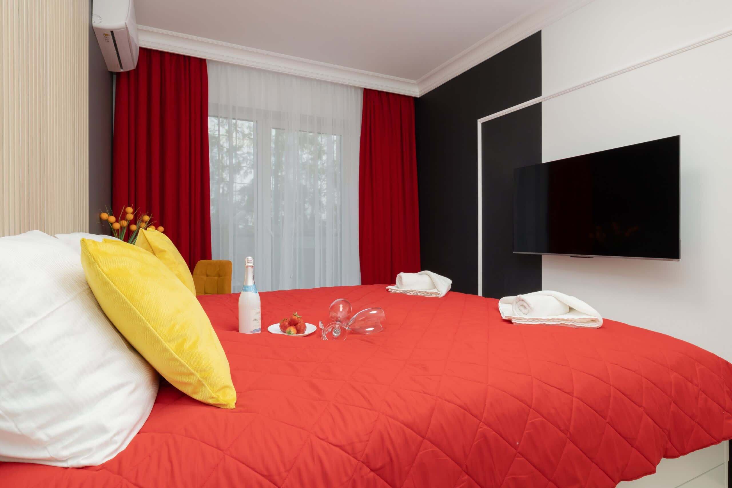 A hotel room with a red bedspread, a flat-screen TV mounted on the wall, a bottle of water, a plate with strawberries, and a telephone.
