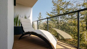 A balcony with a white cushioned chair, a wooden floor, and a view of trees through a glass railing. - Prestigious Apartment | Jacuzzi and Balcony (Dziwnów)